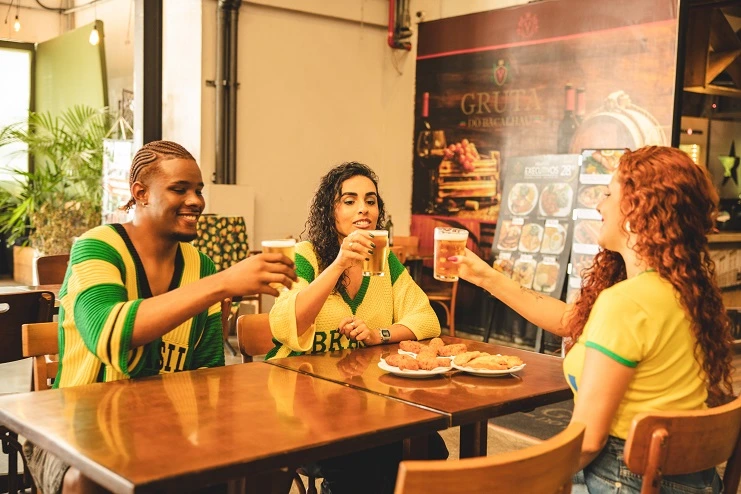 Três amigos brindando em bar com cervejas e petiscos