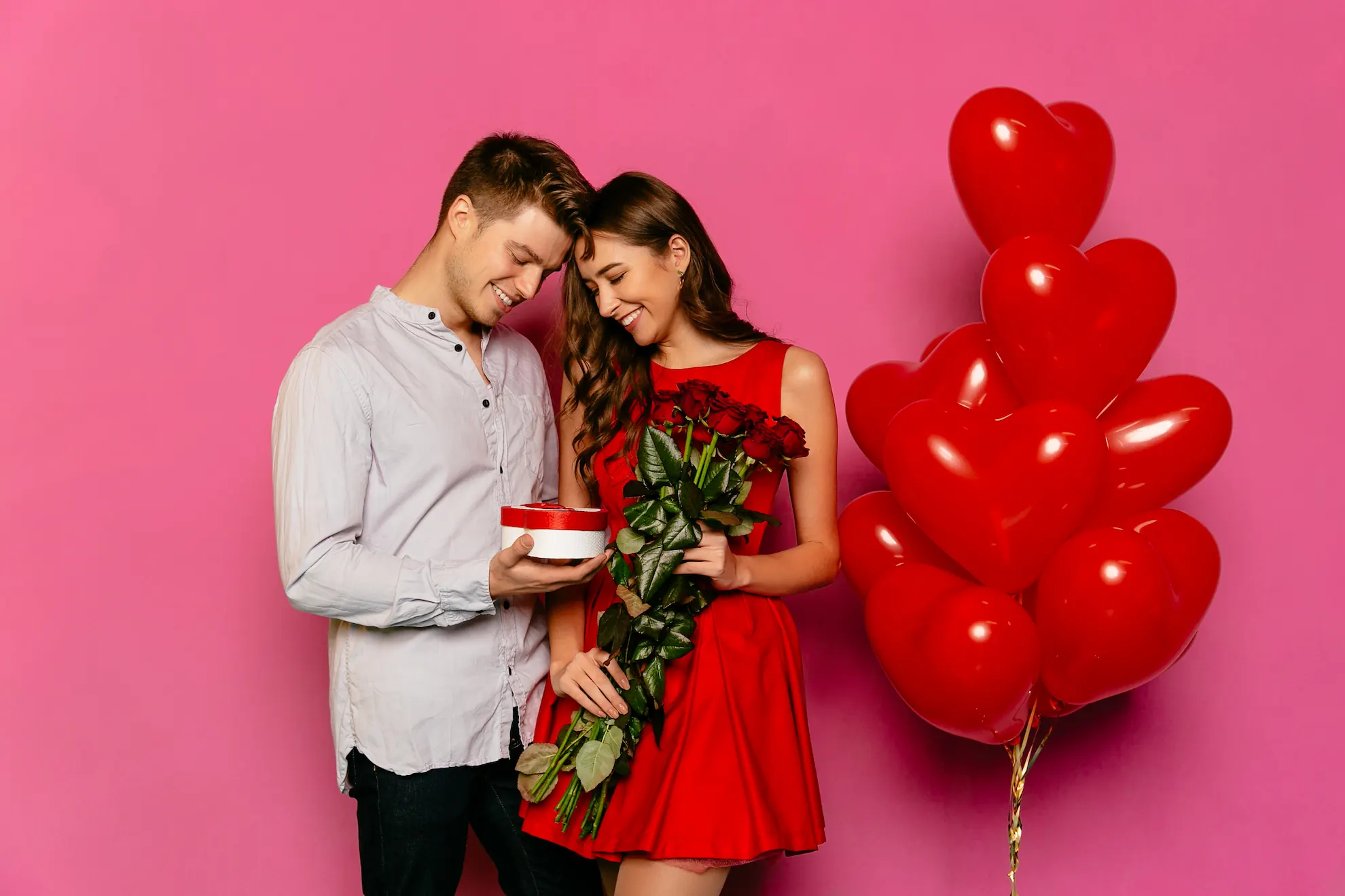 Homem entrega presente e buquê de rosas a mulher em vestido vermelho, cercados por balões de coração em fundo rosa.