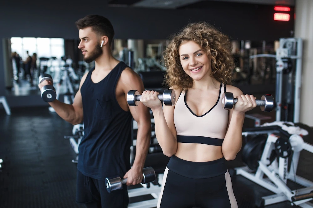 Casal sorridente praticando musculação com halteres dentro de uma academia moderna durante treino.