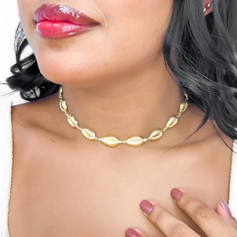 Close no pescoço de uma mulher usando uma gargantilha choker dourada com pingentes em formato de búzios.