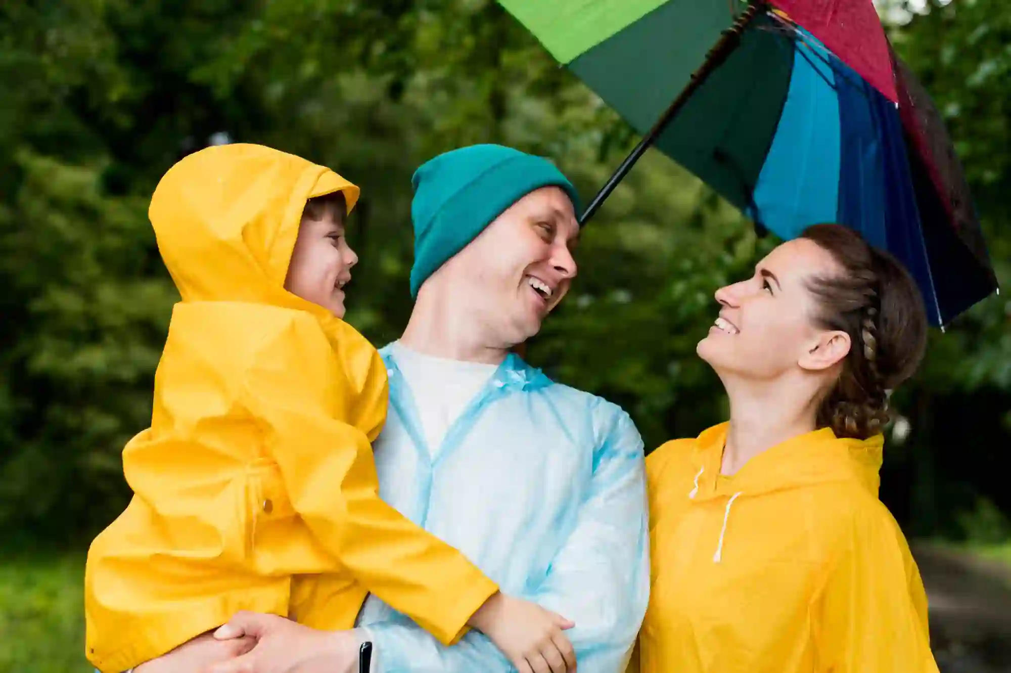 Família de três pessoas a sorrir ao ar livre. O homem, de impermeável azul e gorro verde, segura uma criança vestida com um impermeável amarelo com o capuz colocado. Ao lado, uma mulher também de impermeável amarelo segura um guarda-chuva colorido, tendo como fundo árvores verdes desfocadas.