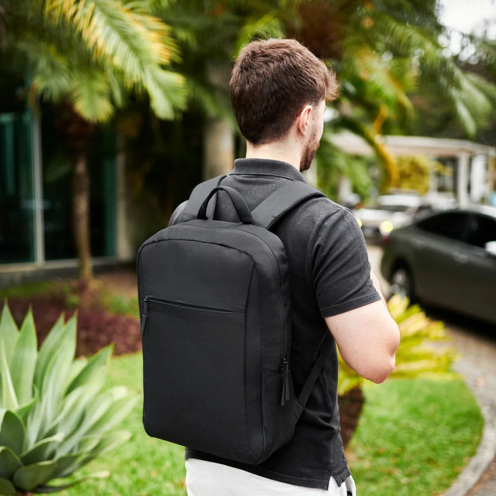 Homem de costas caminhando ao ar livre usando uma mochila executiva preta para notebook em ambiente com árvores.