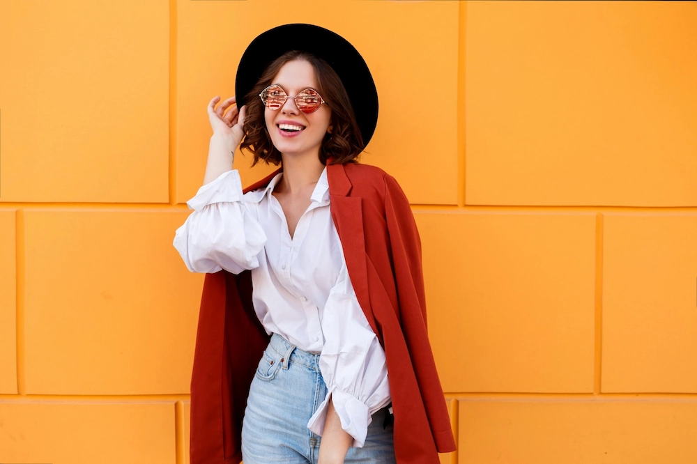 Mulher sorridente usando óculos escuros, chapéu preto e blazer laranja jogado nos ombros contra parede amarela.