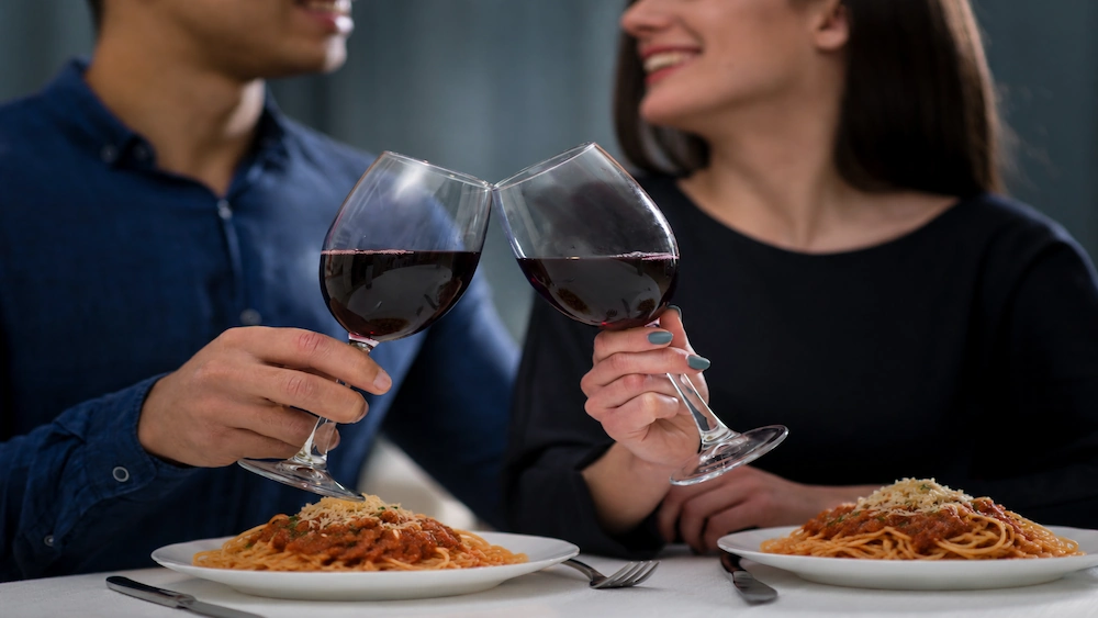 Close em casal brindando com taças de vinho tinto sobre pratos de espaguete à bolonhesa em jantar romântico.