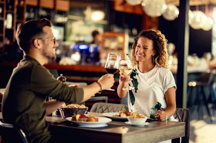 Casal sorridente brinda com taças de vinho tinto e branco durante almoço em restaurante moderno iluminado.