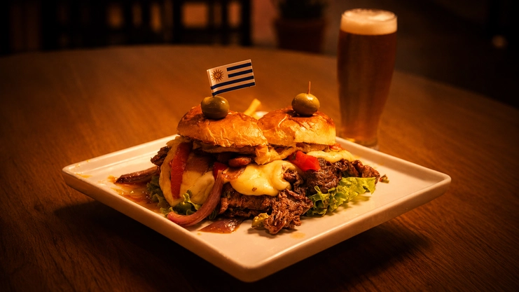 Sanduíche chivito uruguaio aberto com carne, muito queijo e uma bandeirinha do Uruguai ao lado de um copo de chopp.
