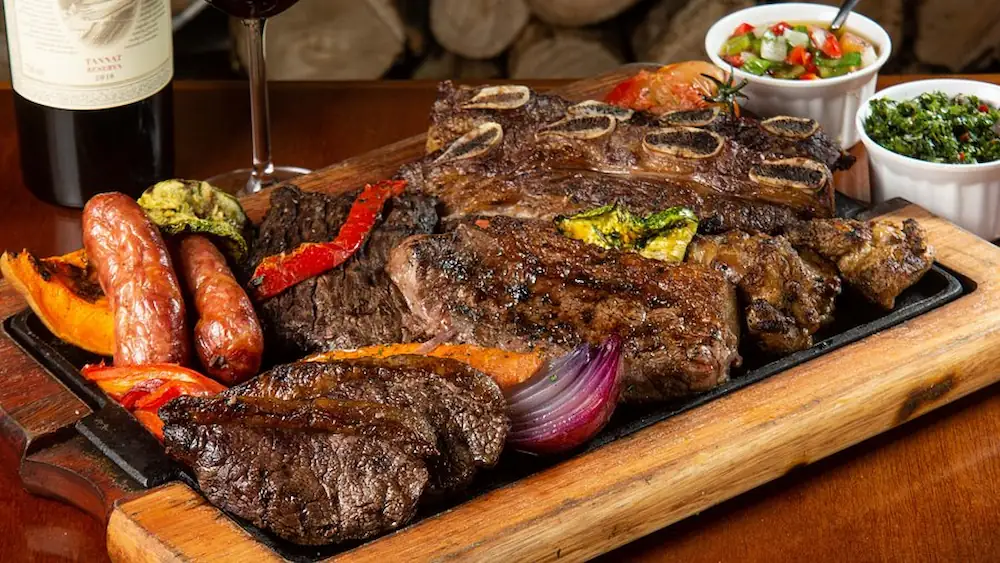 Diversos cortes de carne grelhada, linguiça, vegetais assados, chimichurri e vinagrete em uma tábua de madeira rústica.