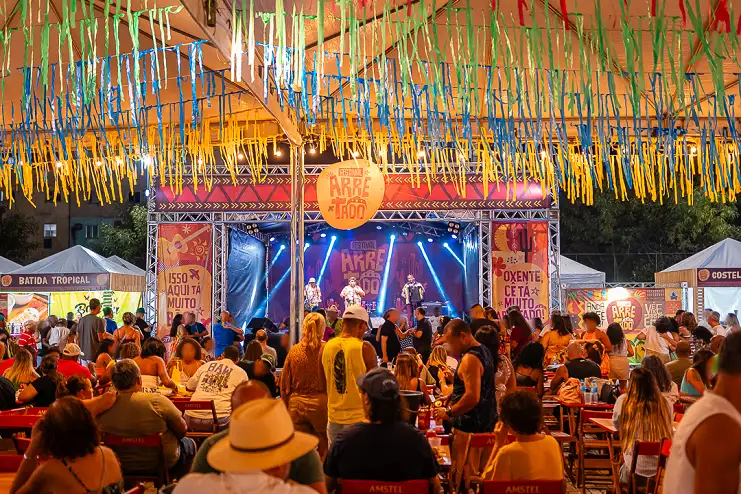 Palco iluminado do Festival Arretado sob fitas coloridas com público em mesas ao ar livre no Uptown Barra, RJ.