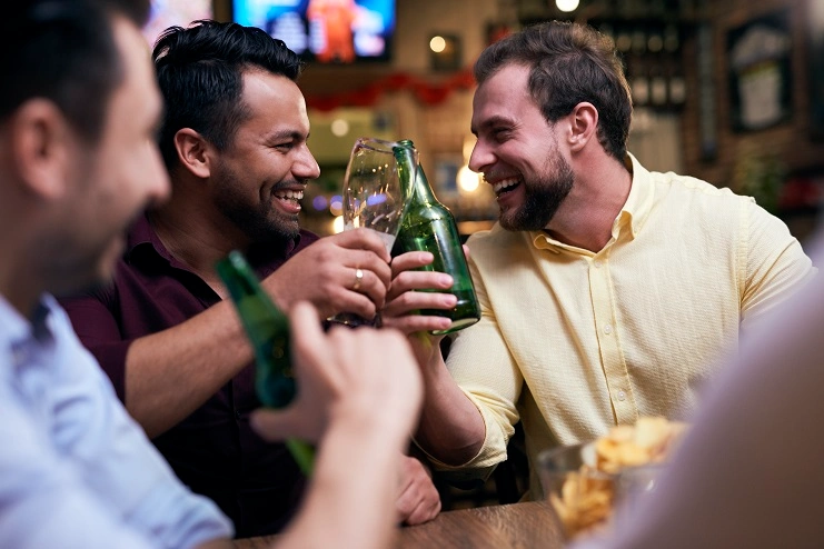 Grupo de amigos homens sorrindo e brindando com garrafas verdes de cerveja em uma mesa de pub animado.