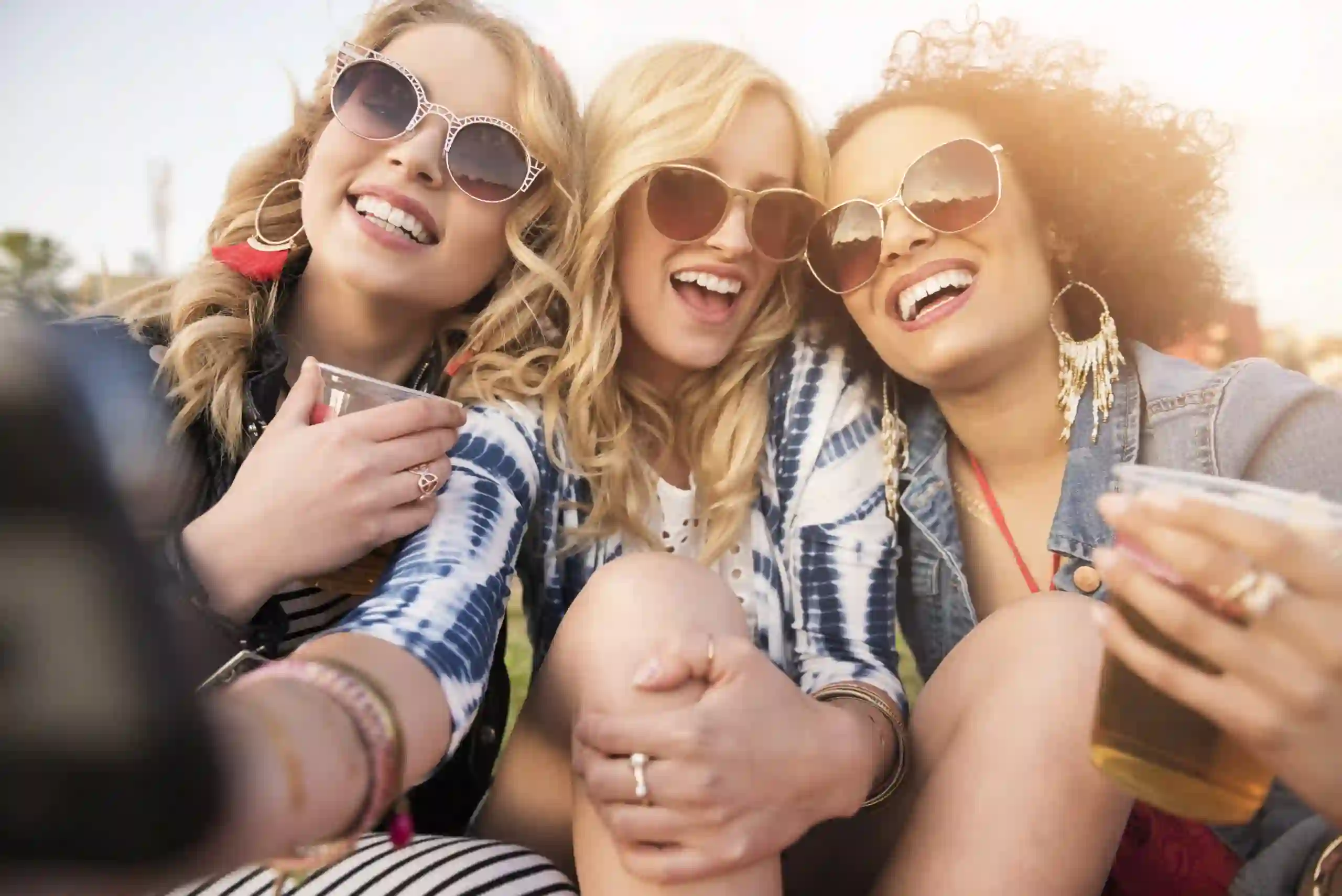 Três mulheres a sorrir enquanto tiram uma selfie ao ar livre sob a luz do sol. Elas usam óculos de sol, joias e roupas casuais, e duas delas seguram copos de plástico com bebida.