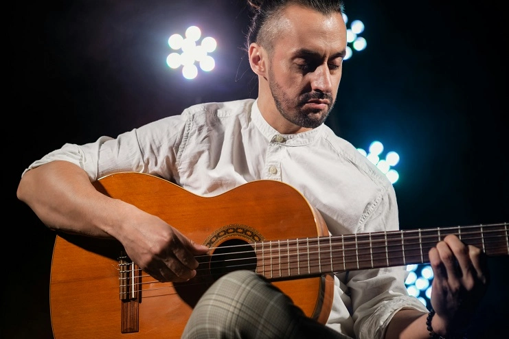 Homem tocando violão acústico concentrado no palco com iluminação cenográfica circular brilhante ao fundo.