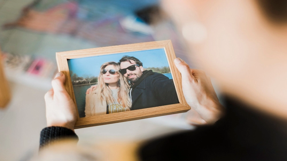 Close nas mãos de alguém segurando um porta-retrato de madeira com uma foto de selfie de casal usando óculos de sol.