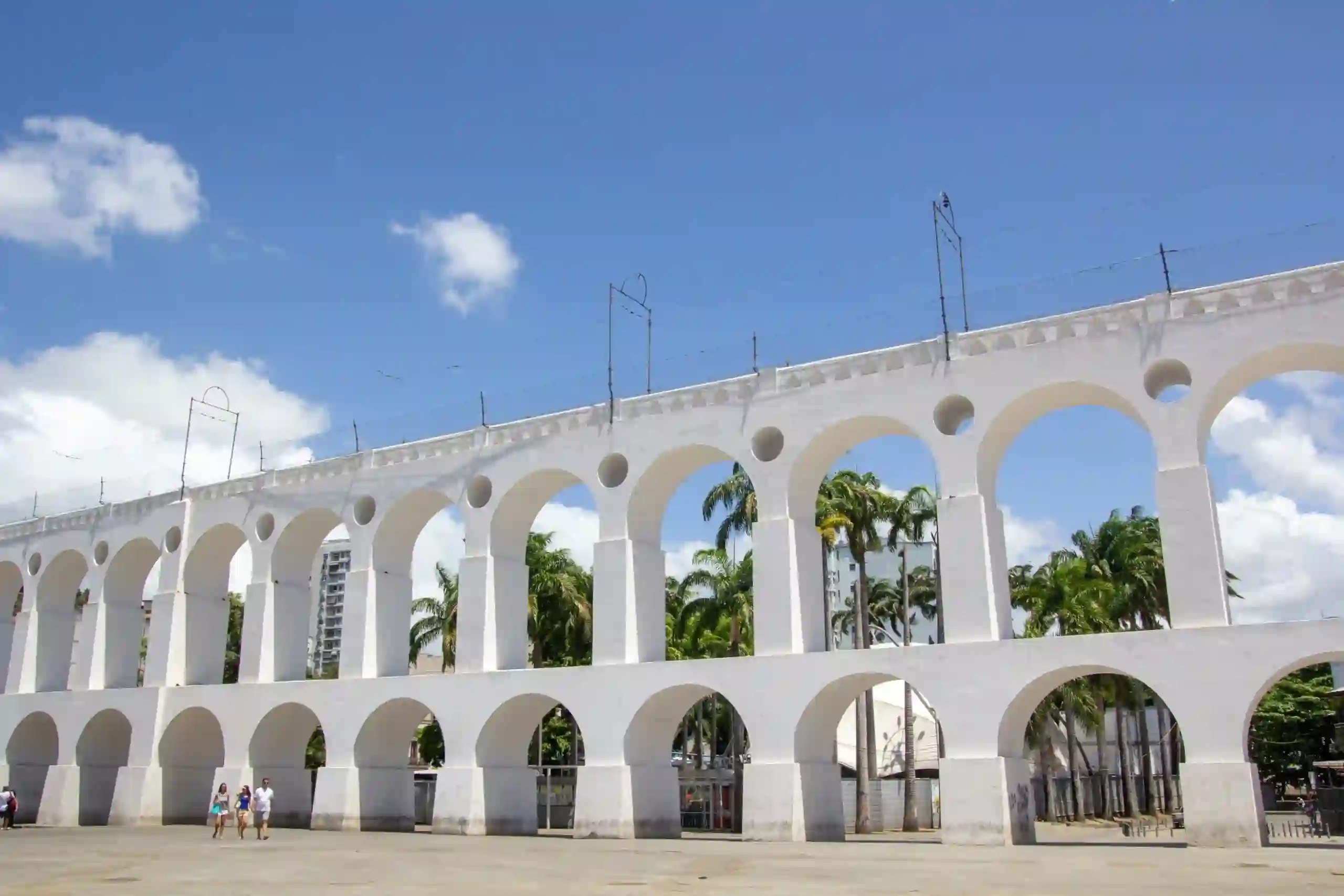 Estrutura branca dos Arcos da Lapa com duas fileiras de arcos sobrepostos. Ao fundo, veem-se palmeiras sob um céu azul com nuvens e, em primeiro plano, algumas pessoas a caminhar numa praça ampla.