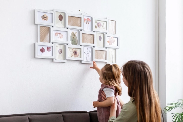 Mãe segura filha pequena no colo apontando para grande painel de porta-retratos brancos com fotos de flores na parede.