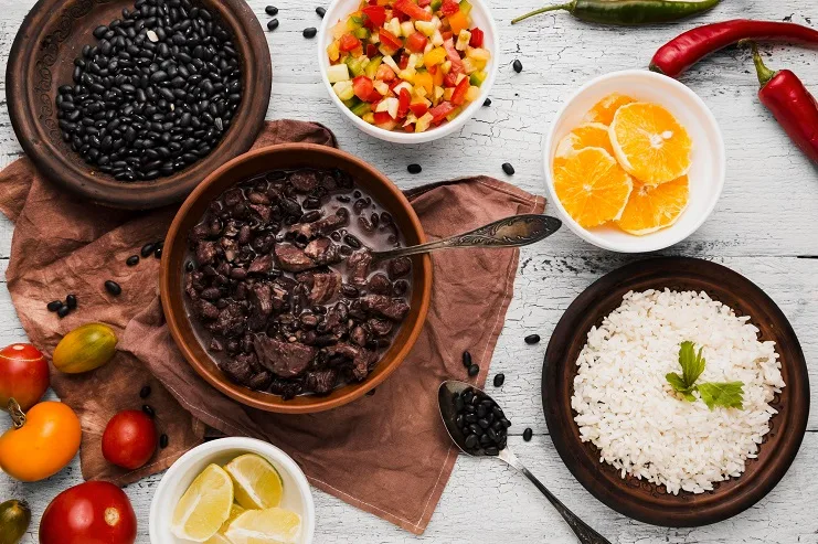 Prato de feijoada em tigela de barro cercado por porções de arroz, vinagrete, laranja e feijão preto cru.