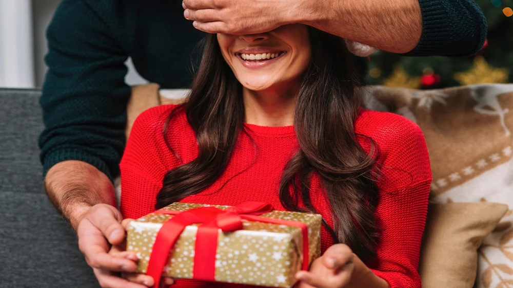 Homem cobre os olhos de mulher sorridente vestindo suéter vermelho para lhe dar um presente de Natal embrulhado.
