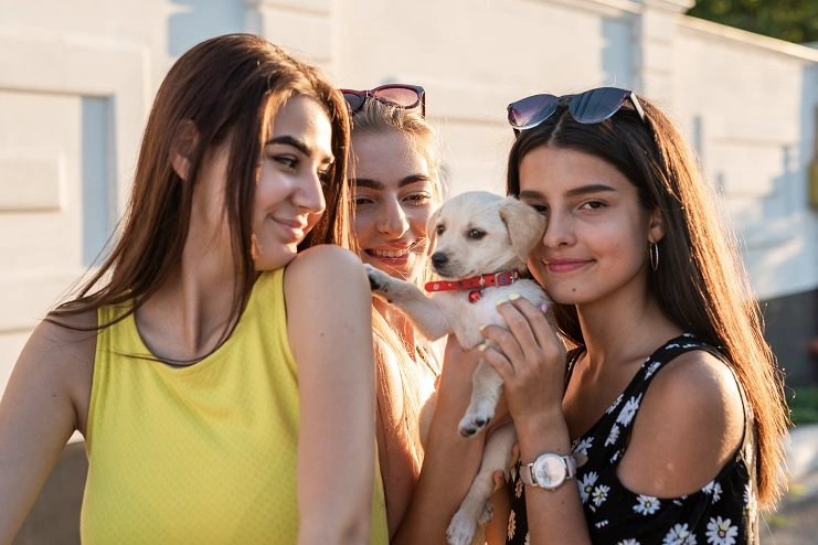 Três jovens sorridentes seguram um pequeno filhote de cachorro branco com coleira vermelha sob a luz do sol.