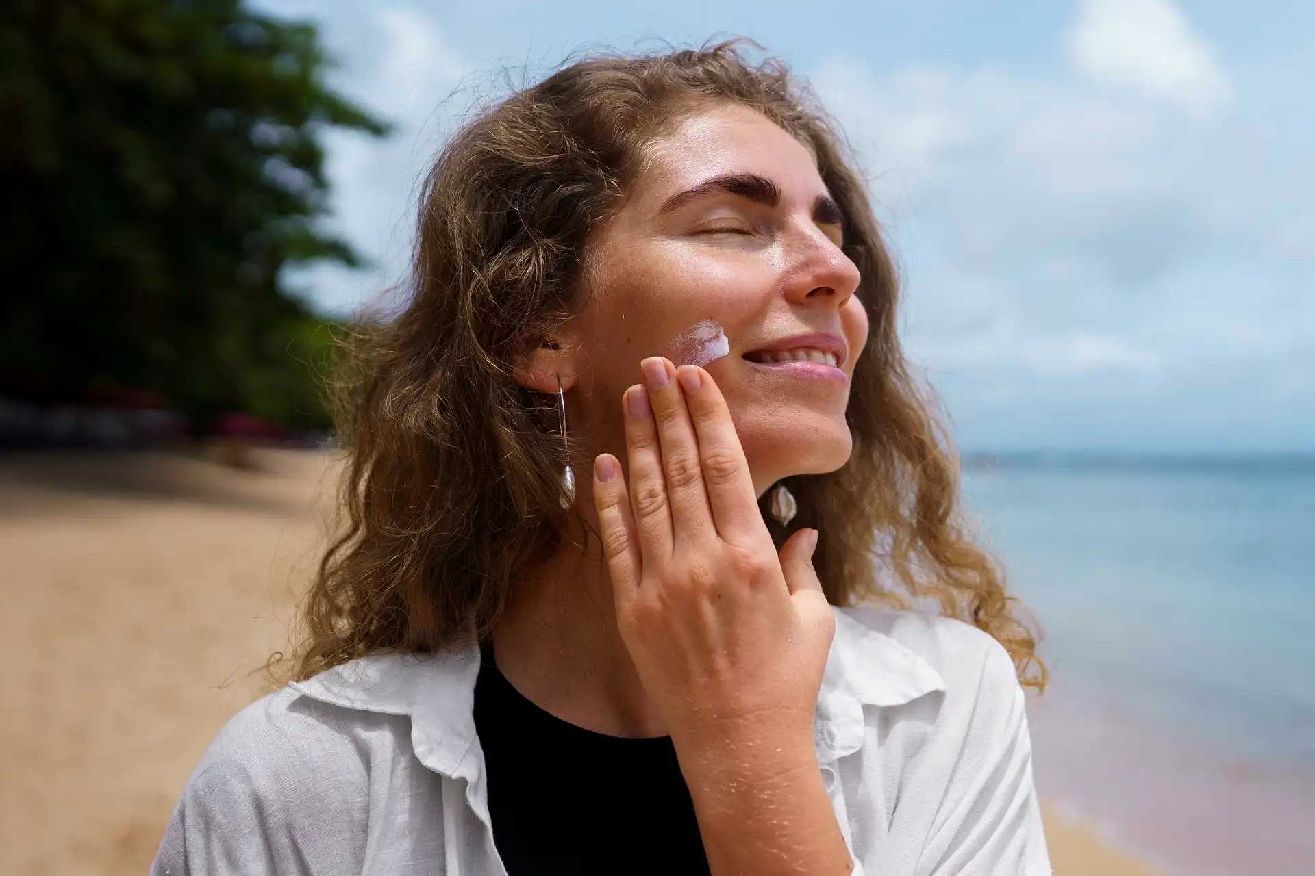 Mulher sorridente de olhos fechados espalha protetor solar na bochecha, em uma praia com mar e árvores ao fundo.
