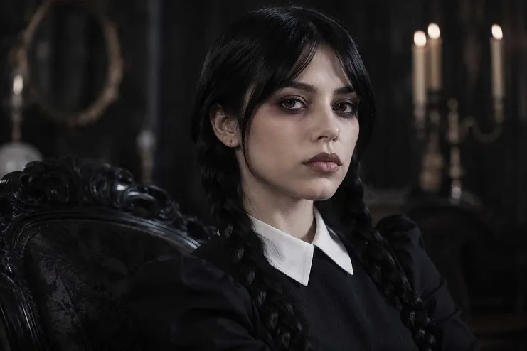 Wandinha Addams encarando a câmera com duas tranças no cabelo, vestido preto e gola branca pontuda.