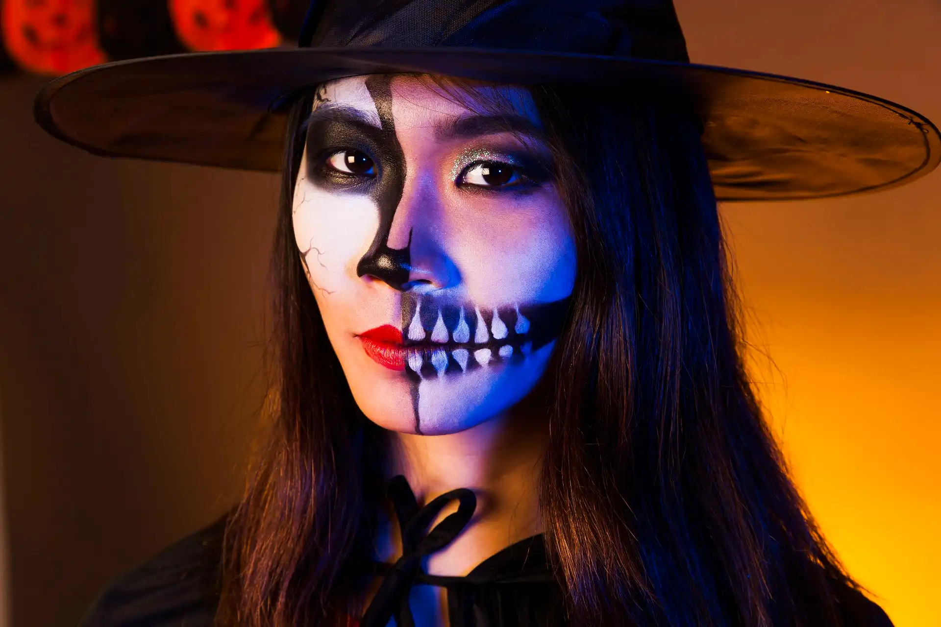 Mulher com chapéu de bruxa e maquiagem artística de caveira dividida metade no rosto.