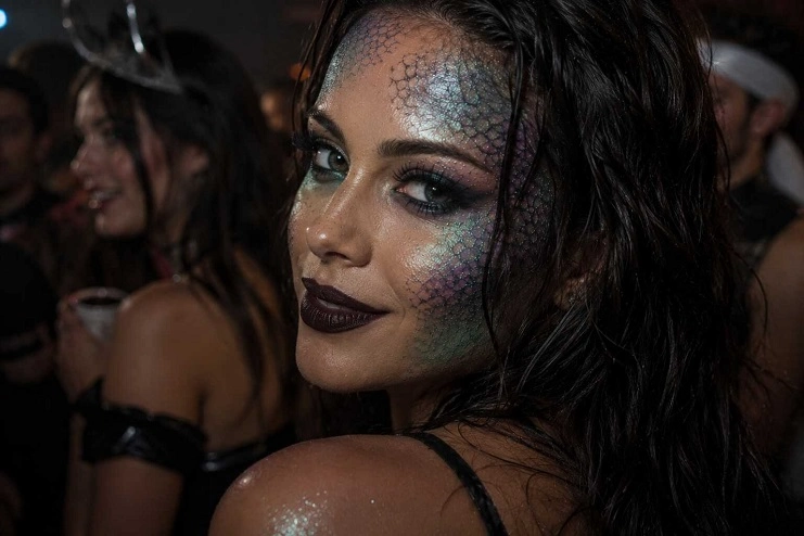 Mulher sorri com maquiagem artística de sereia em tons cintilantes de verde e roxo simulando escamas.