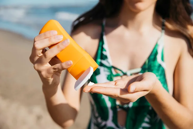 Mulher de biquíni verde aplica creme de protetor solar laranja na palma da mão com a praia ao fundo.